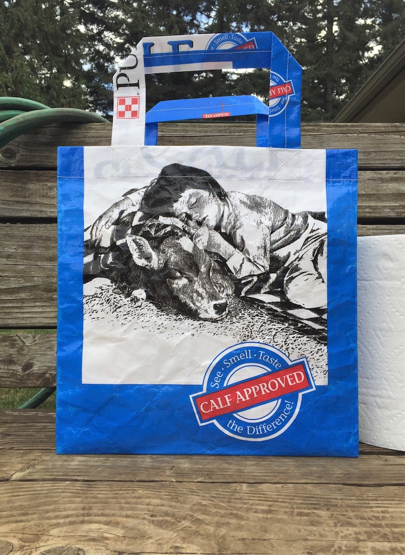 May include: Blue and white tote bag featuring a black and white illustration of a person sleeping next to a calf. The bag has blue handles and the text "CALF APPROVED" in a red and white emblem.