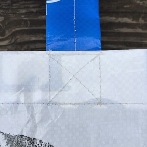 May include: Close-up of a white tote bag with a blue handle. The bag is made of a woven material and has a stitched handle attachment. The handle has white text on a blue background. The bag has a black and white design at the bottom.