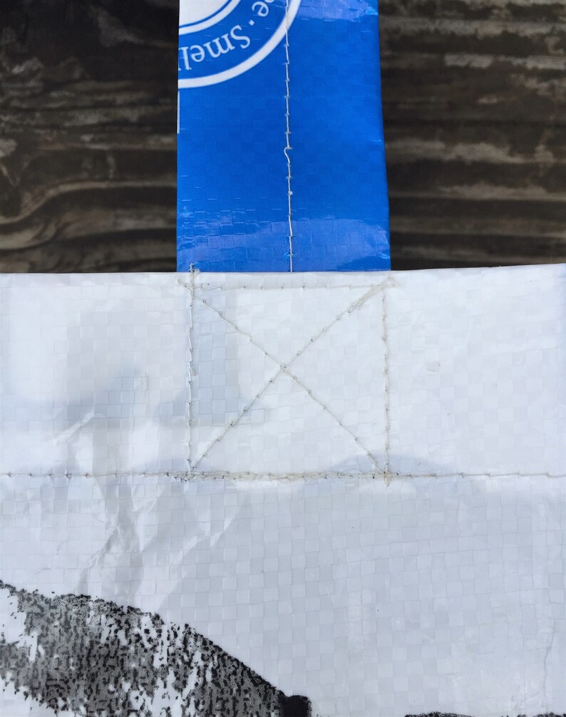 May include: Close-up of a white tote bag with a blue handle. The bag is made of a woven material and has a stitched handle attachment. The handle has white text on a blue background. The bag has a black and white design at the bottom.