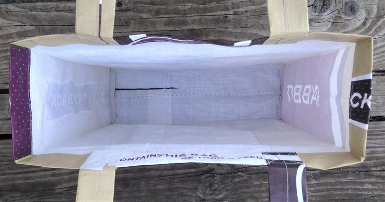 May include: An open, rectangular tote bag with a light-colored interior and a brown exterior. The bag has handles and reinforced corners. The interior is white, and the exterior has text and a purple polka-dot pattern.