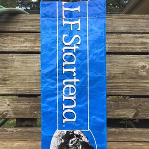 May include: A blue tote bag with white text that reads "LF Startena". The bag has a graphic of a cow's face at the bottom. The bag has blue handles and is sitting on a wooden surface.