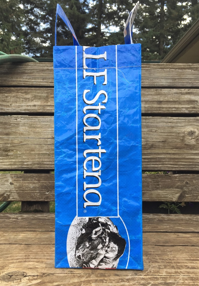 May include: A blue tote bag with white text that reads "LF Startena". The bag has a graphic of a cow's face at the bottom. The bag has blue handles and is sitting on a wooden surface.