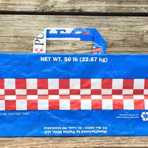May include: Blue and red tote bag with a checkered pattern and a handle. The bag has the text "NET WT. 50 lb (22.67 kg)" and the Purina logo. The bag is made from recycled materials.