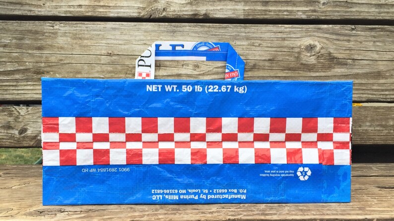 May include: Blue and red tote bag with a checkered pattern and a handle. The bag has the text "NET WT. 50 lb (22.67 kg)" and the Purina logo. The bag is made from recycled materials.
