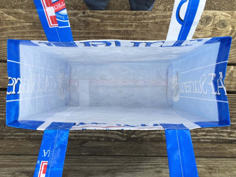 May include: A blue and white tote bag with blue handles. The bag is made from a recycled material and has text printed on the sides. The bag is empty and viewed from the inside.