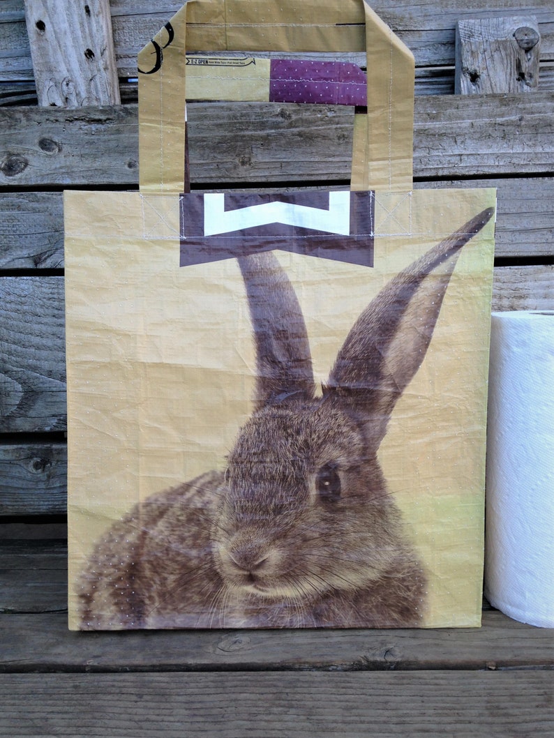 May include: A beige tote bag featuring a detailed illustration of a rabbit's face. The bag has a rectangular shape with a handle and a white and black design at the top. The background is a wooden surface.