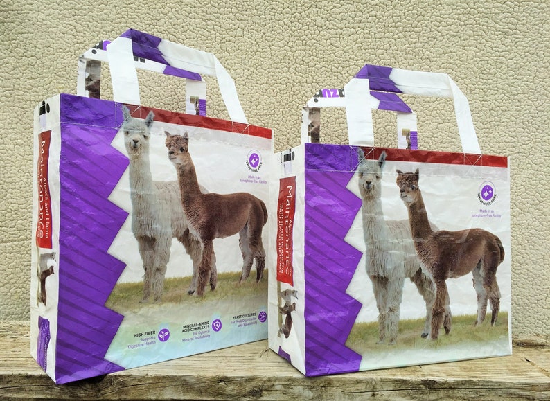 May include: Two tote bags with images of alpacas. The bags are white with purple and red accents. The bags have handles and feature the words "Maintenance" and "Alpaca and Llama". The bags also have text about high fiber and mineral content.