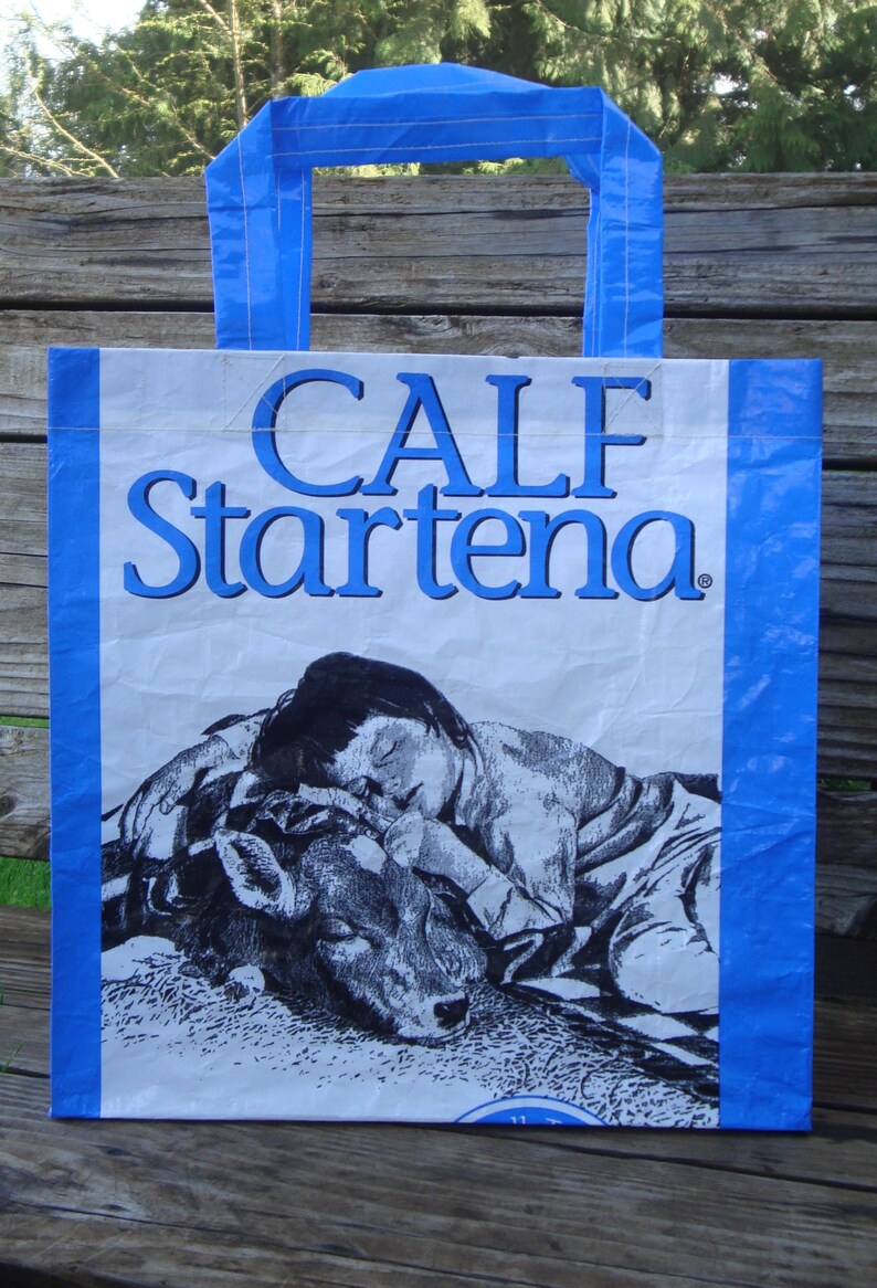 May include: Blue and white tote bag with the words "CALF Startena" in blue. The bag features a black and white illustration of a person sleeping with a calf. The bag has blue handles.