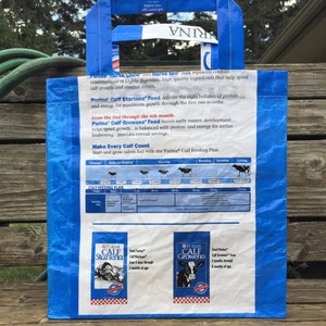 May include: A blue and white Purina Calf Startena Feed bag with handles. The bag features text and graphics about calf nutrition and growth. The bag is made of a durable, possibly recycled material, and is designed for carrying.