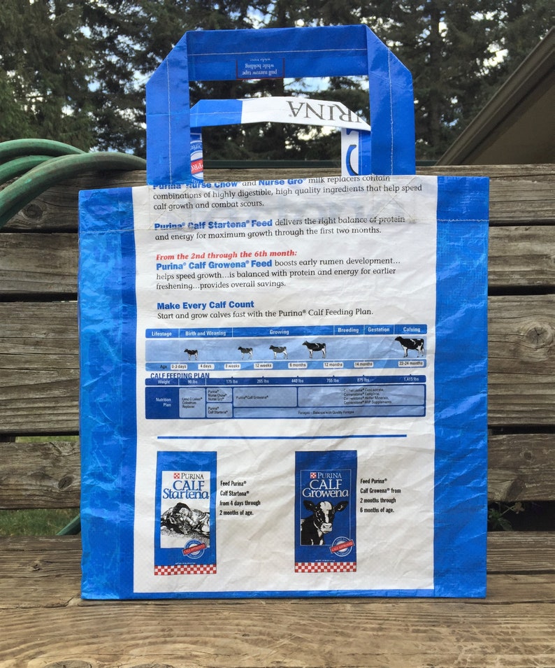 May include: A blue and white Purina Calf Startena Feed bag with handles. The bag features text and graphics about calf nutrition and growth. The bag is made of a durable, possibly recycled material, and is designed for carrying.