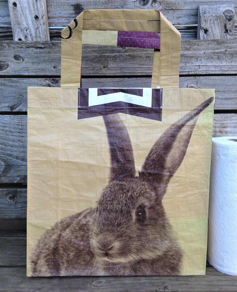 May include: A beige tote bag featuring a detailed illustration of a rabbit. The bag has a rectangular shape with a brown and white chevron design and purple polka dot accents. The handles are made of the same beige material.