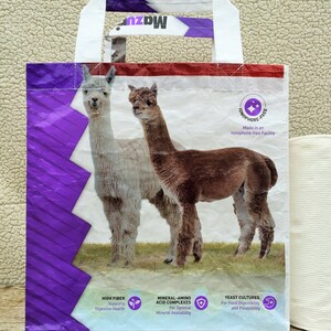 May include: A white and purple tote bag featuring two alpacas on a grassy field. The bag has white handles and the text "Mazuri" at the top. The bag also includes text about high fiber, mineral-amino acid complexes, and yeast cultures.