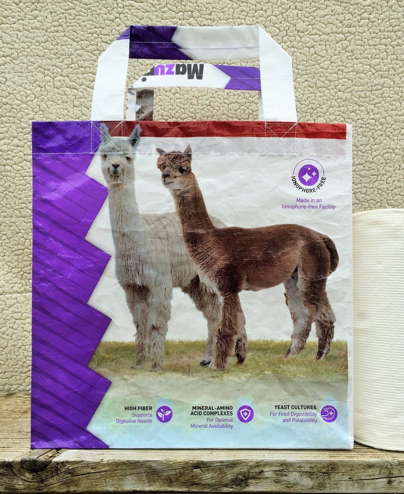 May include: A white and purple tote bag featuring two alpacas on a grassy field. The bag has white handles and the text "Mazuri" at the top. The bag also includes text about high fiber, mineral-amino acid complexes, and yeast cultures.