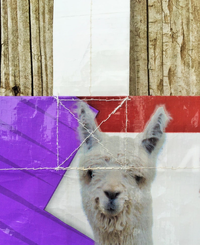 May include: A close-up of a white llama with a smiling expression, set against a backdrop of purple, red, and white panels. The image is stitched together with white thread, and the background includes wooden planks.