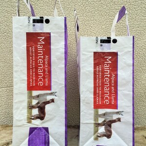 May include: Two white feed bags with purple accents and handles. The bags have the word "Maintenance" in a red rectangle, along with "Alpaca and Llama" and "Fortified Feed". The bags feature illustrations of alpacas.
