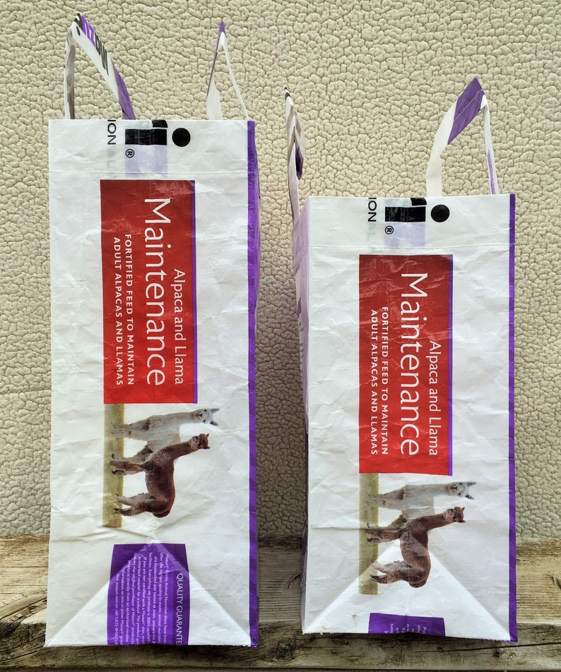 May include: Two white feed bags with purple accents and handles. The bags have the word "Maintenance" in a red rectangle, along with "Alpaca and Llama" and "Fortified Feed". The bags feature illustrations of alpacas.