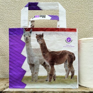 May include: A white and purple tote bag featuring two alpacas on a green field. The bag has white handles and a red stripe at the top. The text "Mazuri" is visible on the handle. The bag is labeled "Ionophore-free".
