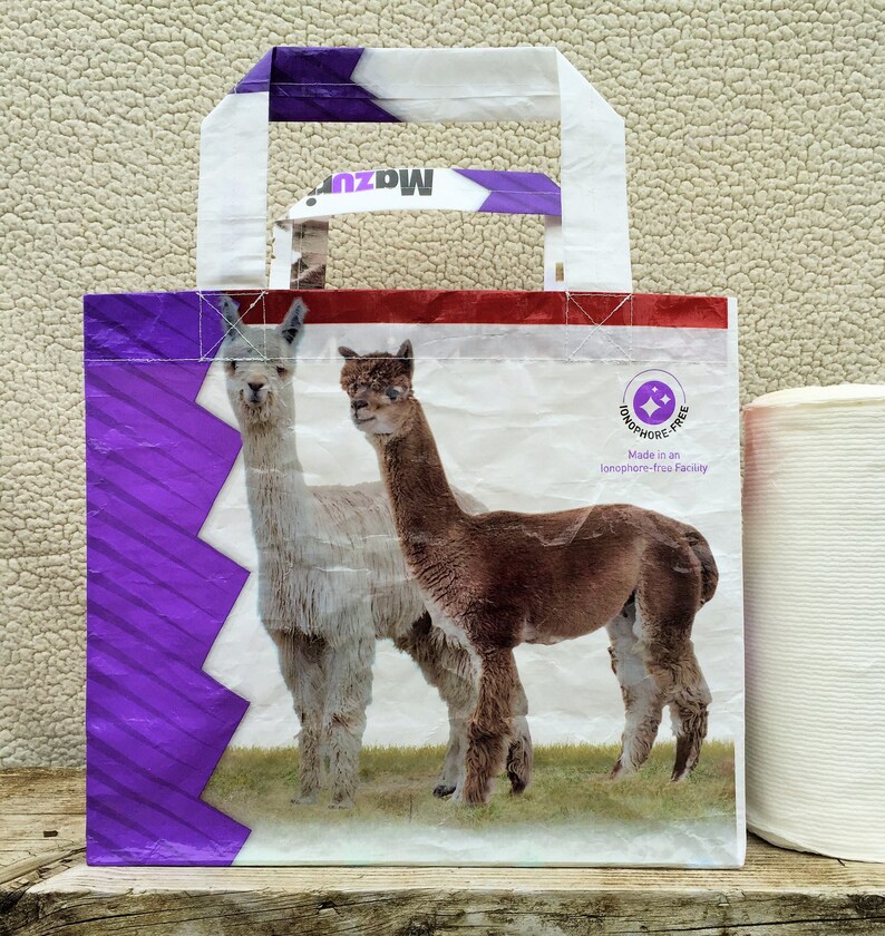 May include: A white and purple tote bag featuring two alpacas on a green field. The bag has white handles and a red stripe at the top. The text "Mazuri" is visible on the handle. The bag is labeled "Ionophore-free".
