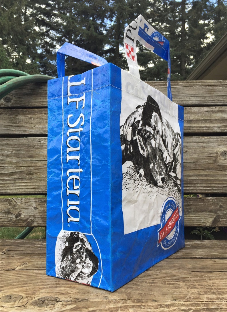 May include: A blue and white tote bag made from recycled material. The bag features a black and white image of a cow and the text "LF Star Tenda". The bag has handles for carrying.