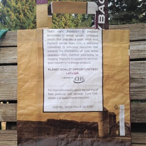 May include: A brown paper tote bag with a handle featuring the word "Payback" and text from CHS. The bag has a landscape image of a farm and silos at the bottom. The handle is made of a patterned material.