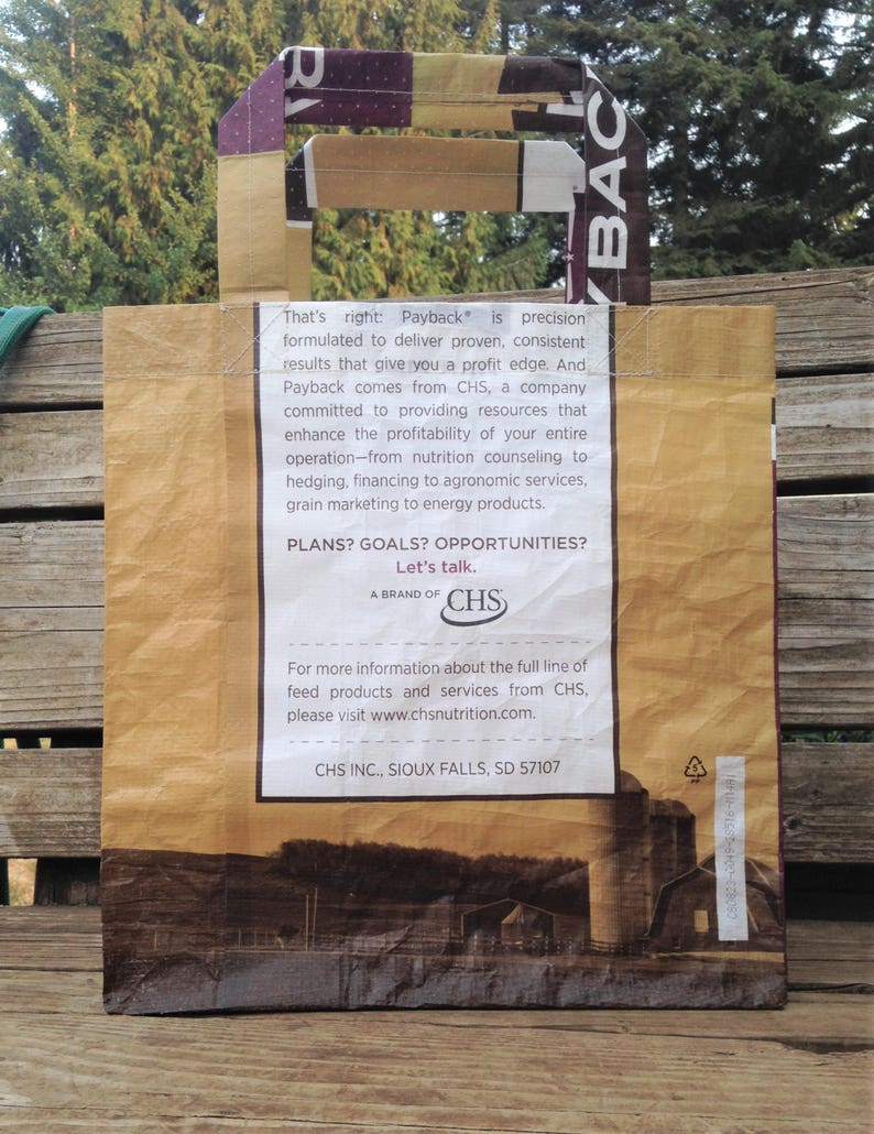 May include: A brown paper tote bag with a handle featuring the word "Payback" and text from CHS. The bag has a landscape image of a farm and silos at the bottom. The handle is made of a patterned material.