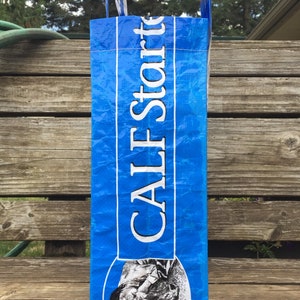 May include: A blue bag with the text "CALF Starter" in white. The bag has a picture of a calf on the bottom. The bag is sitting on a wooden surface.