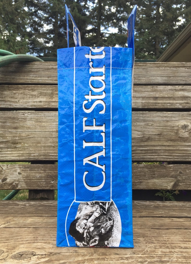 May include: A blue bag with the text "CALF Starter" in white. The bag has a picture of a calf on the bottom. The bag is sitting on a wooden surface.