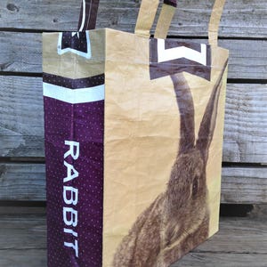 May include: A tote bag with a rabbit illustration. The bag features a large image of a rabbit on a tan background. The side of the bag is purple with white polka dots and the word "RABBIT" in white.