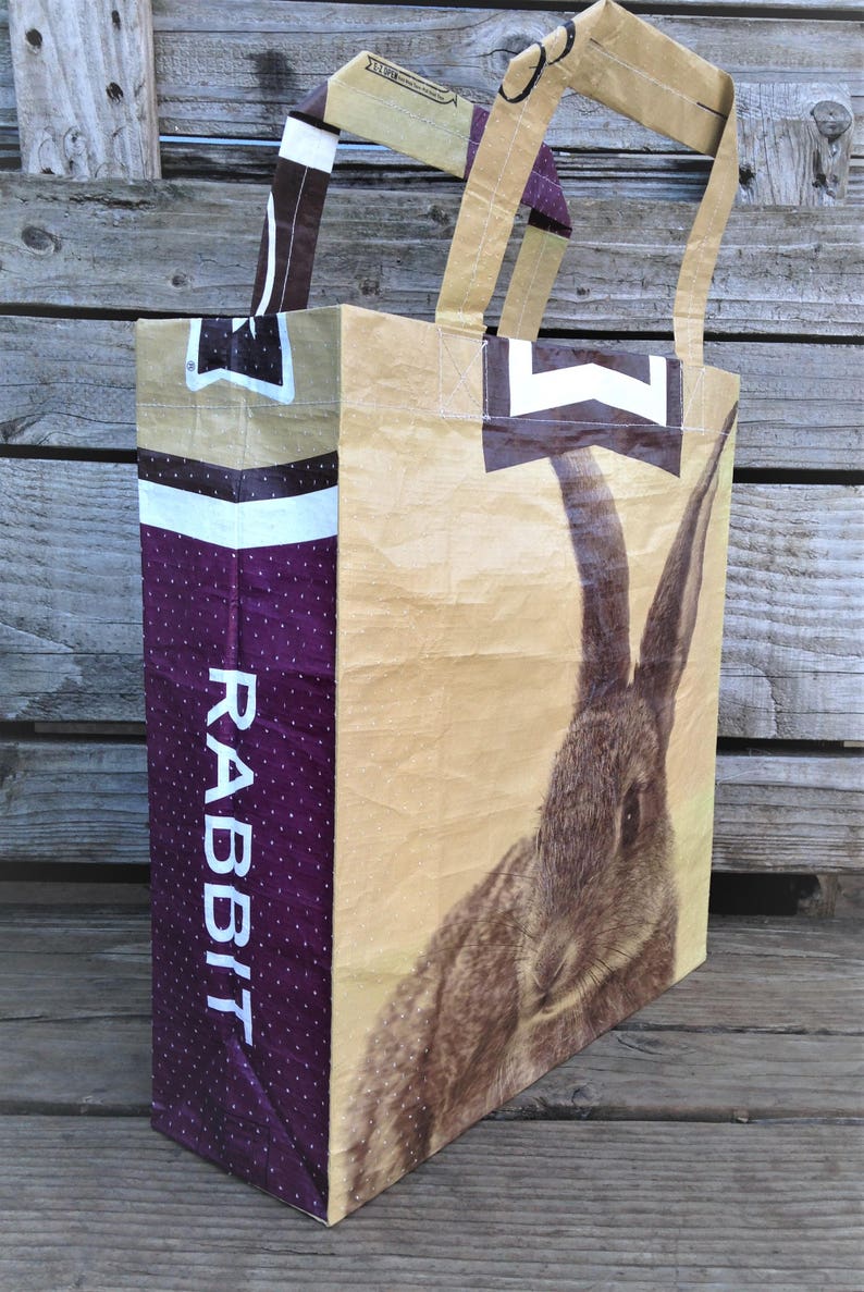 May include: A tote bag with a rabbit illustration. The bag features a large image of a rabbit on a tan background. The side of the bag is purple with white polka dots and the word "RABBIT" in white.