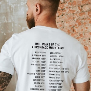 May include: White t-shirt with black text listing the highest peaks of the Adirondack Mountains. The text includes the name of each peak and its elevation in feet. The shirt is worn by a person with a beard and tattoos.