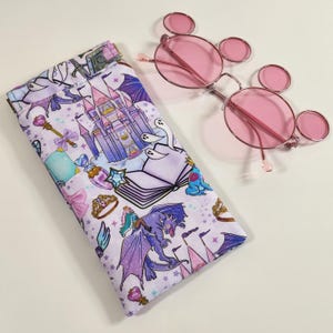 May include: Pink-tinted round sunglasses with a matching patterned case. The case features a whimsical design with a castle, dragon, and other fantasy elements in shades of purple, blue, and pink. The sunglasses have a silver frame.