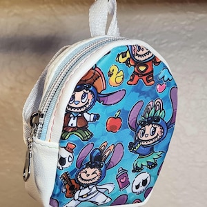 May include: A small, white and blue backpack with a cartoon character print. The backpack features a zipper closure, a small handle, and a silver-colored zipper pull. The print includes various cartoon characters, apples, and skulls.