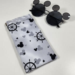 May include: Black sunglasses with circular lenses and small circular side accents, paired with a rectangular fabric pouch. The pouch features a light gray background with a repeating pattern of black ship wheels and solid black mouse-ear silhouettes.