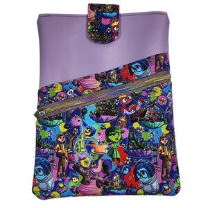 May include: A rectangular pouch featuring a solid light purple upper section and a diagonal lower section with a colorful print of animated characters. The pouch includes a zippered pocket across the front and a snap-button closure at the top.