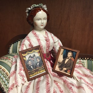 May include: A porcelain doll wearing a white and red striped dress with a green and white floral headpiece. The doll is holding two framed pictures. One picture shows two men in suits and the other shows a man in a suit sitting in a chair.