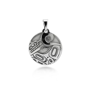 May include: A circular silver pendant featuring an engraved stylised animal design with deep black recessed lines. The metal surface has a polished finish, and the piece includes a small silver bail at the top for attachment to a chain or cord.