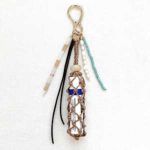 May include: A handcrafted lip balm holder with a macramé design in brown, featuring a silver clasp and decorative beads in white, beige, and turquoise. The holder is designed to carry a standard-sized lip balm tube.