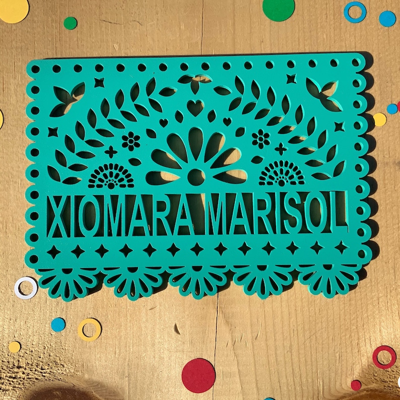 May include: A teal blue paper banner with a traditional Mexican design. The banner has cutouts of flowers, leaves, and stars. The name "XIOMARA MARISOL" is written in white letters across the center of the banner.