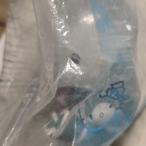 May include: A clear plastic bag containing a light blue item with a Hello Kitty design. The bag is partially transparent, and the contents are slightly obscured. The Hello Kitty design is visible in blue.