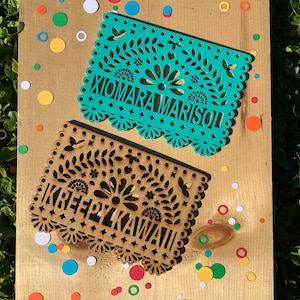 May include: Two paper banners with intricate cut-out designs. The top banner is teal blue with the text "XIOMARA MARISOL". The bottom banner is brown with the text "KREEPY KAWAII".