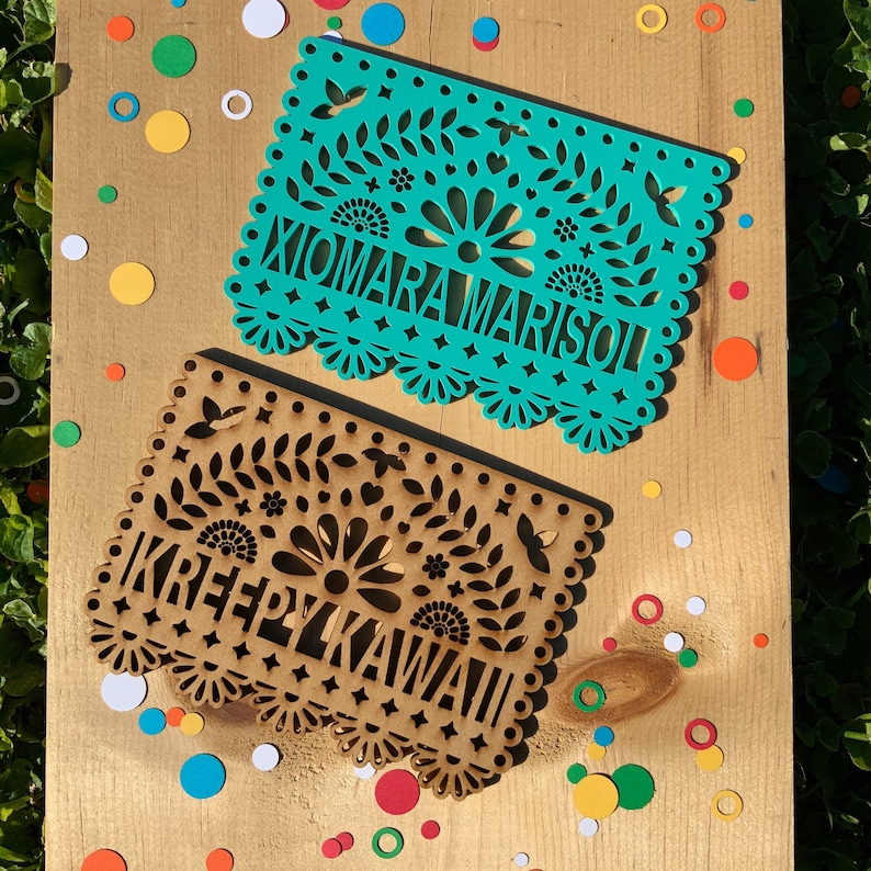 May include: Two paper banners with intricate cut-out designs. The top banner is teal blue with the text "XIOMARA MARISOL". The bottom banner is brown with the text "KREEPY KAWAII".