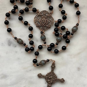 May include: A rosary featuring black beads strung on copper-colored wire. The rosary includes a decorative centerpiece and a detailed cross pendant. The beads are round and evenly spaced, creating a symmetrical design.