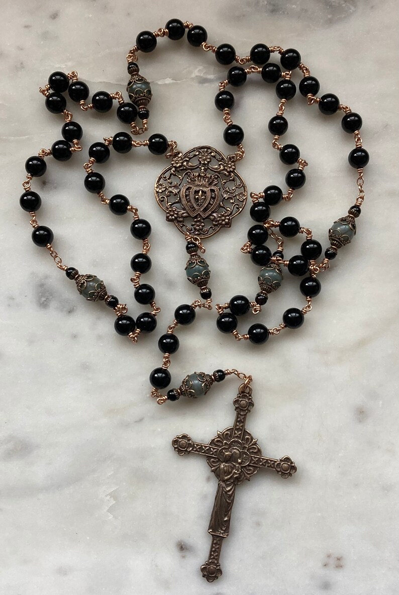 May include: A rosary featuring black beads strung on copper-colored wire. The rosary includes a decorative centerpiece and a detailed cross pendant. The beads are round and evenly spaced, creating a symmetrical design.