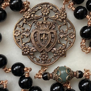 May include: Close-up of a rosary with black beads and copper wire. The centerpiece is an ornate, bronze-colored metal emblem featuring a crown, fleur-de-lis, and floral details. A green bead is also visible.