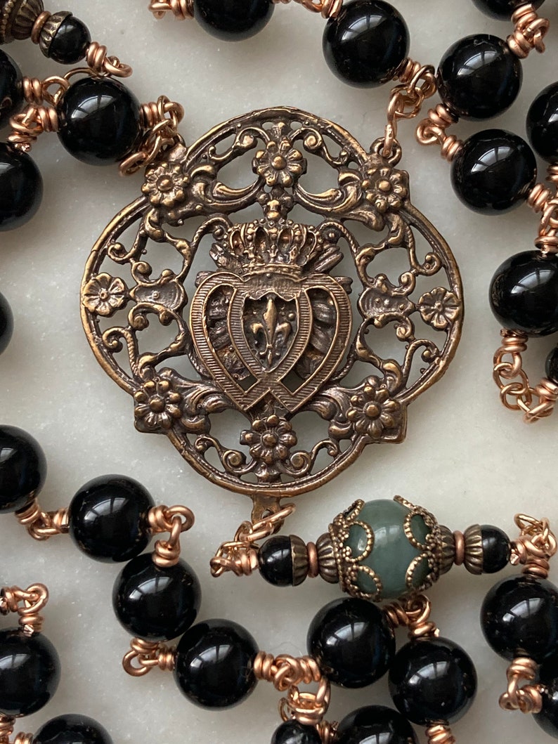 May include: Close-up of a rosary with black beads and copper wire. The centerpiece is an ornate, bronze-colored metal emblem featuring a crown, fleur-de-lis, and floral details. A green bead is also visible.