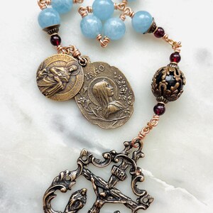 May include: A rosary featuring light blue beads and bronze-toned metal components. The design includes a crucifix, decorative medallions, and a filigree bead. The rosary is a religious item.