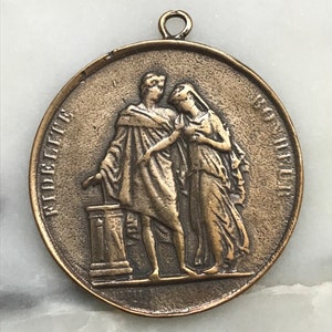 May include: A bronze medal with a raised image of two figures standing side-by-side. The figures are wearing robes and are holding hands. The medal has an inscription around the edge that reads "ST. AUGUSTINE, FL. 1887".
