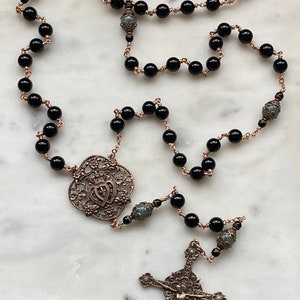 May include: A rosary with black beads and bronze-colored wire. The rosary features a decorative centerpiece and a detailed crucifix. The beads are round and evenly spaced, creating a traditional religious accessory.