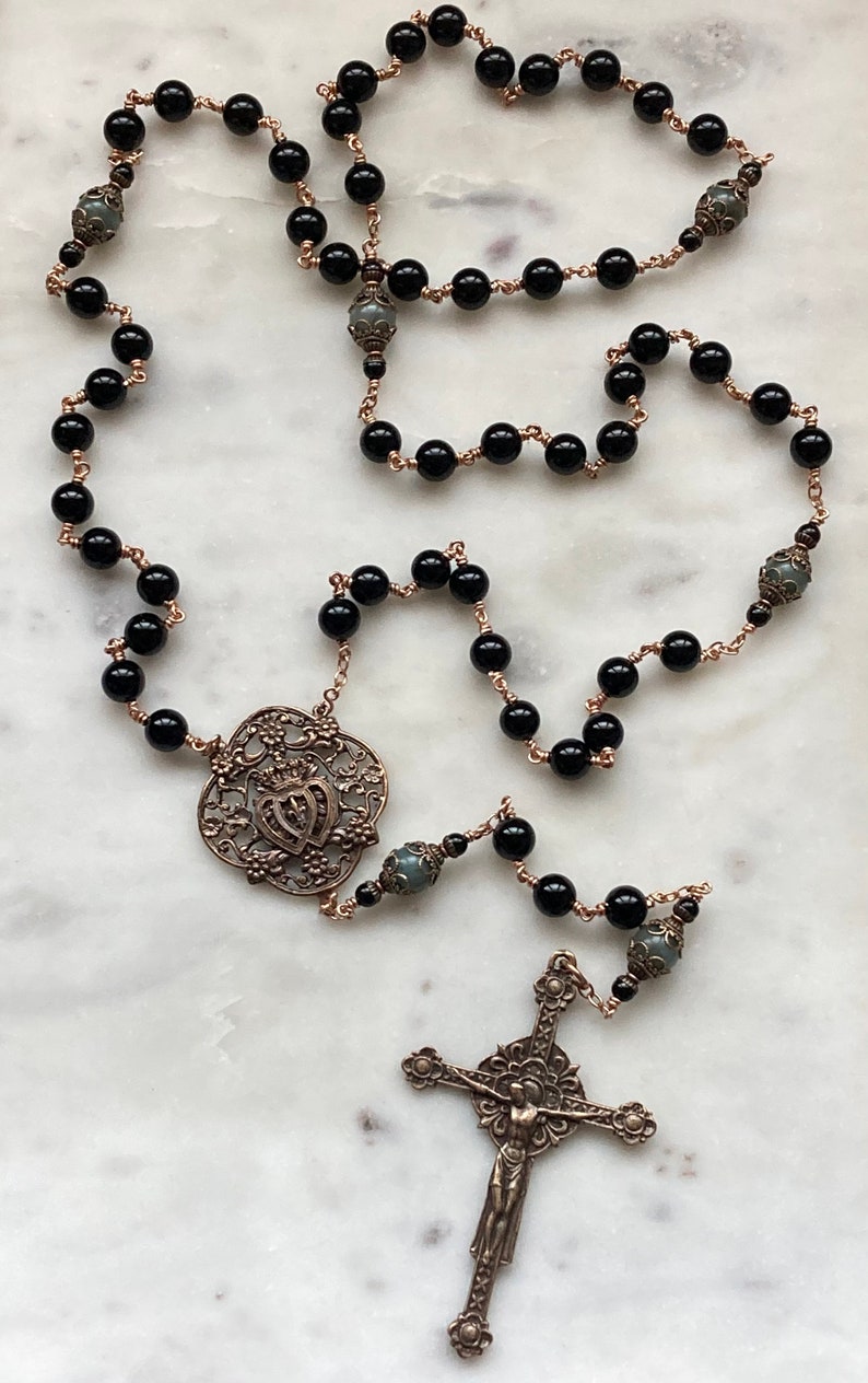 May include: A rosary with black beads and bronze-colored wire. The rosary features a decorative centerpiece and a detailed crucifix. The beads are round and evenly spaced, creating a traditional religious accessory.