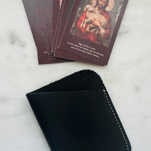 May include: A set of burgundy prayer cards with the text "Surrender Novena Day 1" and an image of Jesus holding a lamb. A black leather card holder is below the cards.